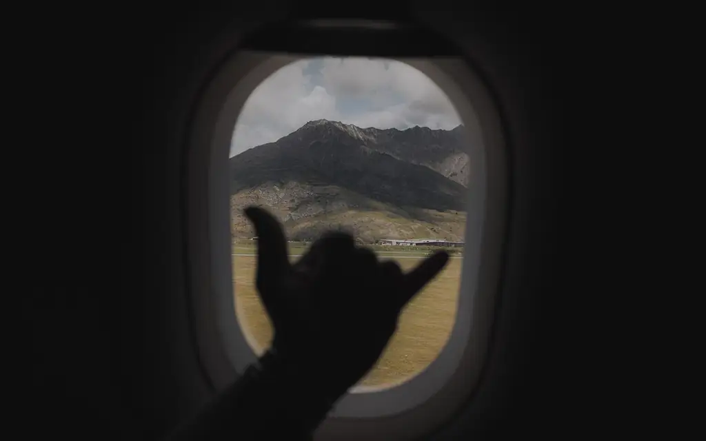 Hand making shaka gesture through airplane window with scenic Queenstown mountains in view