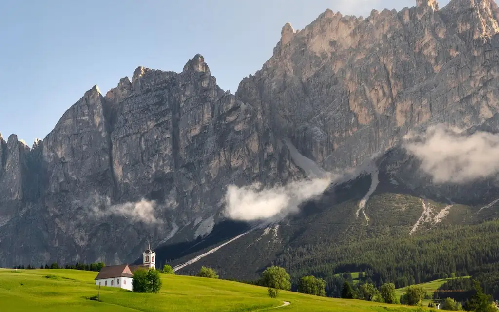 A serene view of a church against the majestic Dolomites mountains with lush greenery and clouds