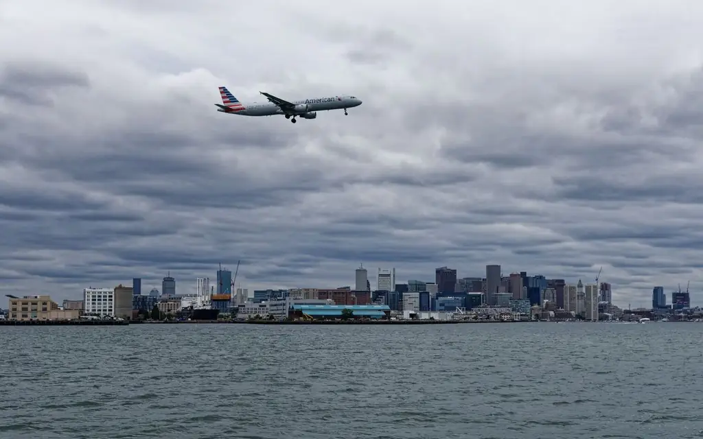 Passenger plane flying over water towards Boston skyline, cloudy day