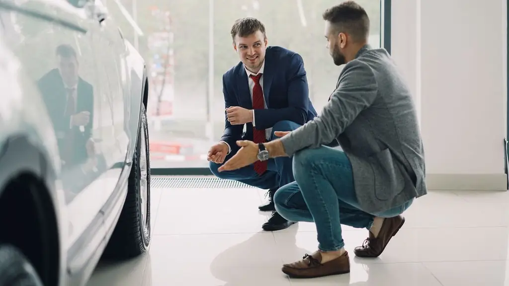 Two men discussing car features in a showroom, kneeling near a vehicle