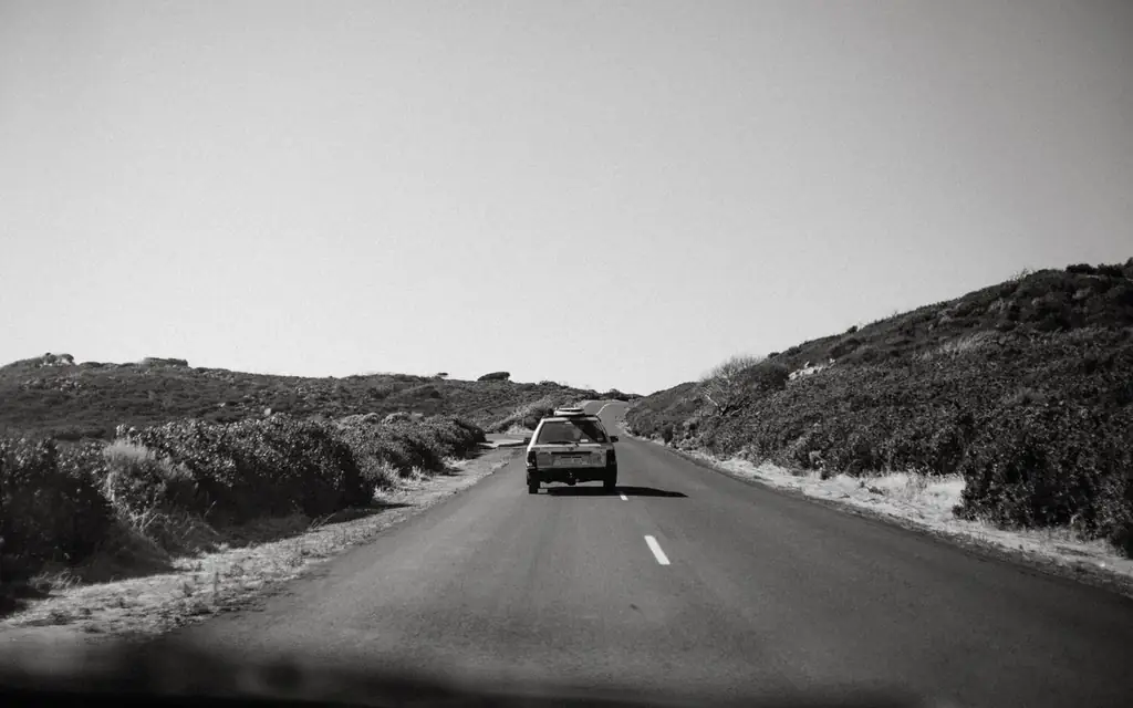 Black and white of automobile driving on empty asphalt highway along green hills in countryside against cloudless sky in nature