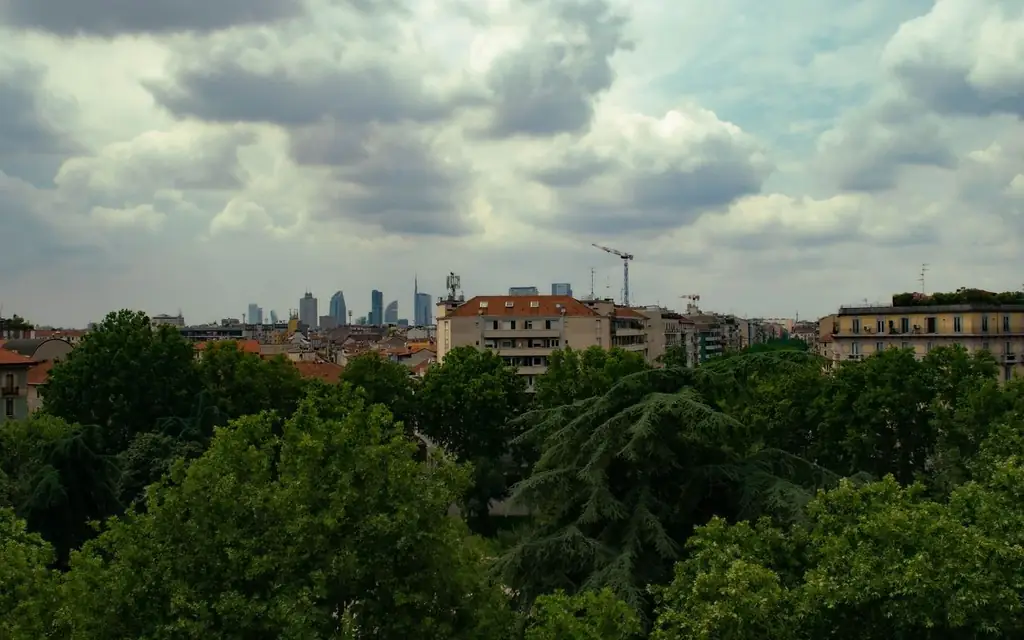 Skyline of Milan with clear view of urban buildings amidst lush green trees under a cloudy sky