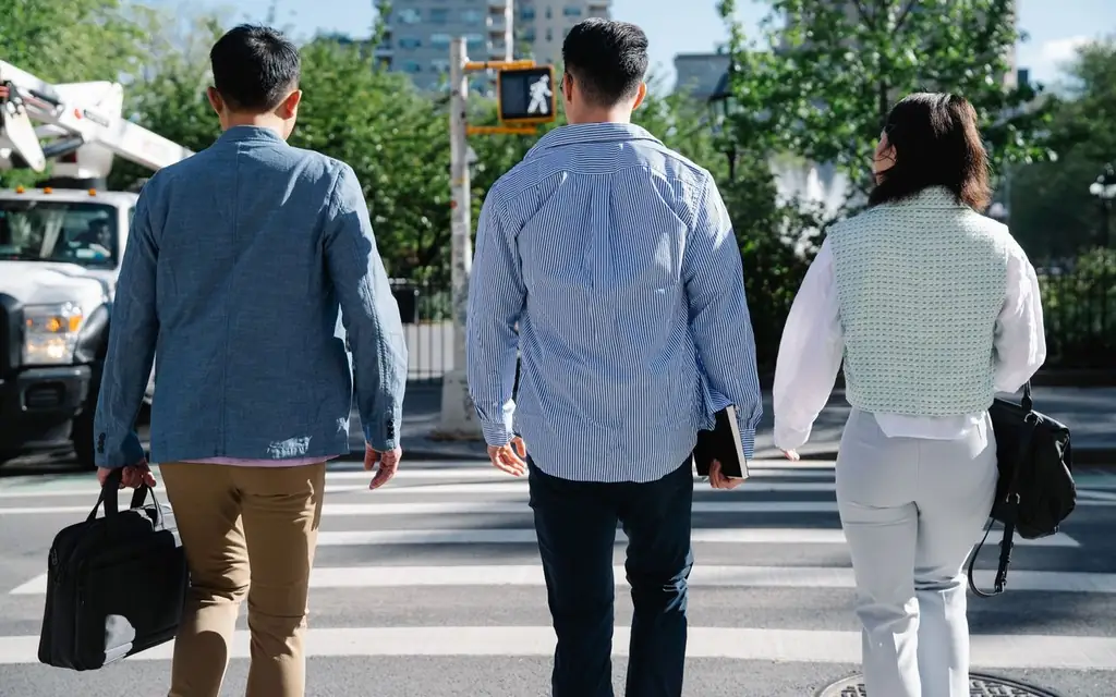 Three adults crossing a city road at a pedestrian crossing, carrying handbags under a clear sky